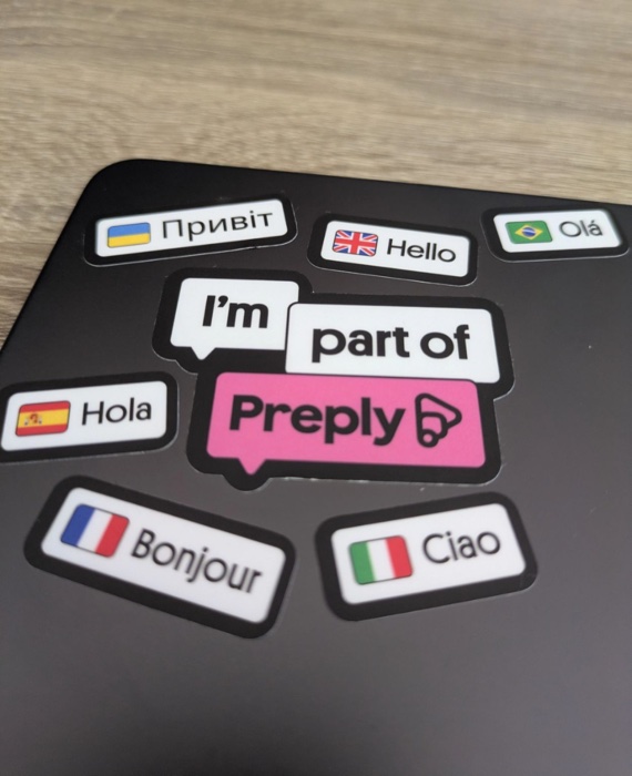 Stickers on a dark laptop lid: “I'm part of Preply” in the middle, surrounded by greetings in different languages — Привіт, Hello, Olá, Hola, Bonjour, Ciao.